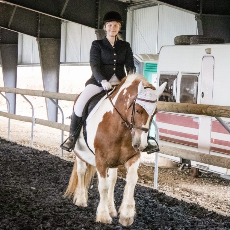 Cavalière en situation de handicap en tenue d'équitation sur un cheval pie dans une carrière intérieure.